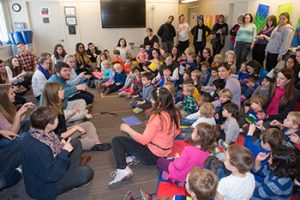 Music education students sitting on the floor leading a large group of preschool students sitting on the floor, as faculty and teachers watch standing in the back.