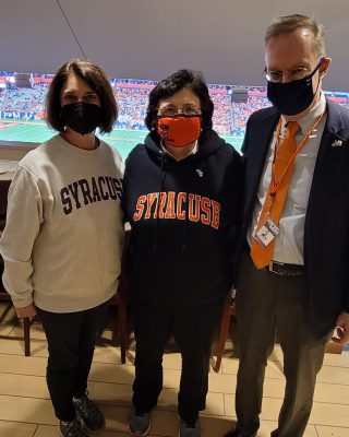 three people in syracuse clothing pose for the camera with a football field in the background