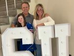 kate, david, and amy allyn smile at the camera standing behind large light up S U letters.