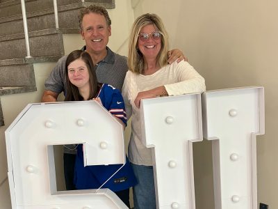 kate, david, and amy allyn smile at the camera standing behind large light up S U letters.
