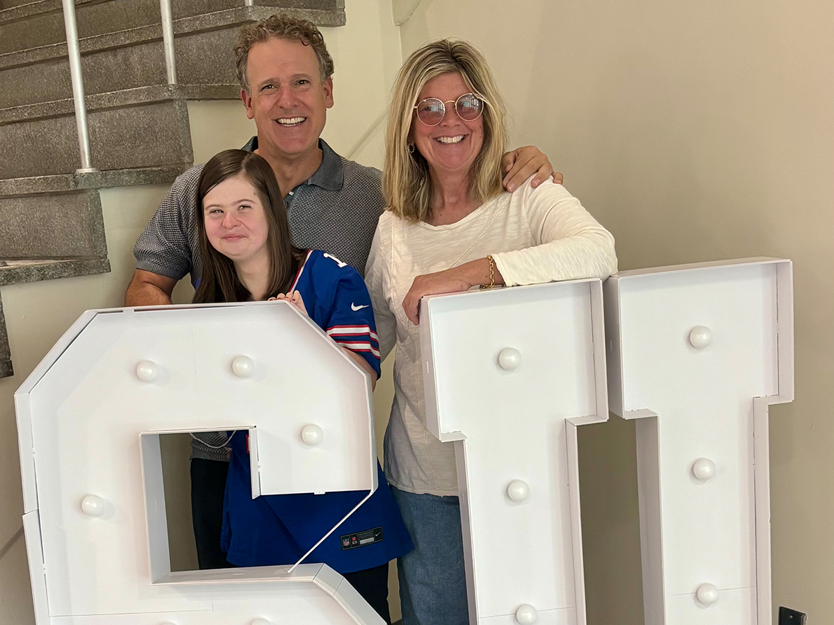 kate, david, and amy allyn smile at the camera standing behind large light up S U letters.