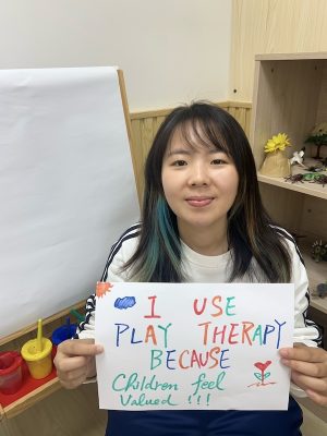 Coco Wing Yun Wong holds a sign thats says "I use play therapy because children fveel valued"