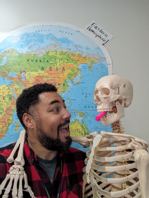 A male teacher stands next to a skeleton, with a highlighter in its mouth