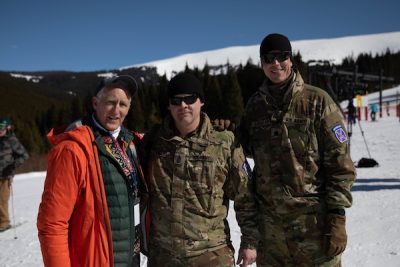One civilian and two military members stand on a snowy hill