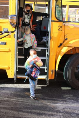 an elementary school student in a mask gets off a yellow school bus
