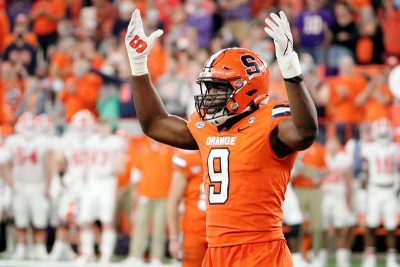 Kingsley Jonathan in an orange football uniform on the field, with both hands raised and a crowd visible behind him