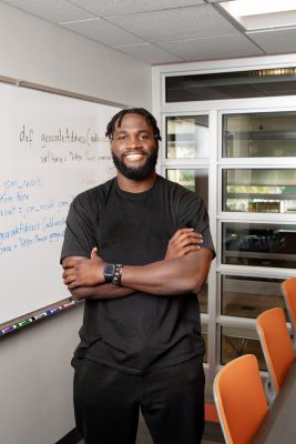 Portrait of Kingsley Jonathan standing in a classroom smiling and and with arms folded, a dry erase board behind him